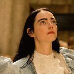 Emma Stone em cena de Pobres Criaturas (2023), vestindo um traje azul com detalhes brancos e olhando para cima em um ambiente clássico.